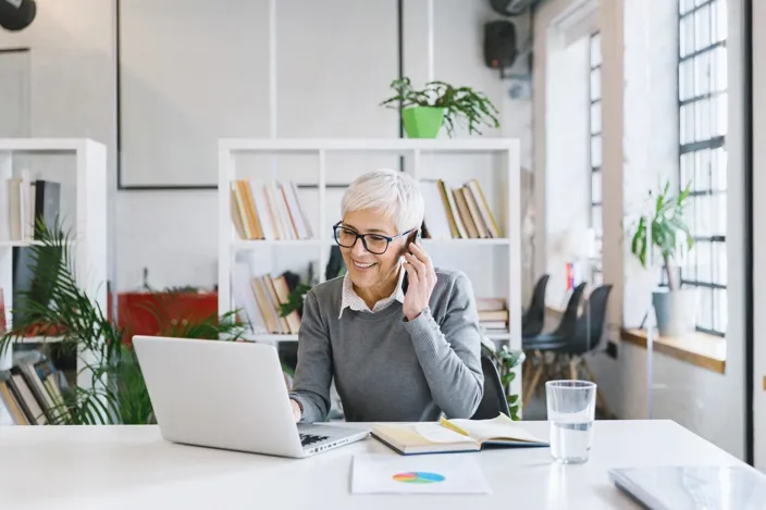 Senior female entrepreneur working in the office and talking on her smartphone, simultaneously checking her laptop