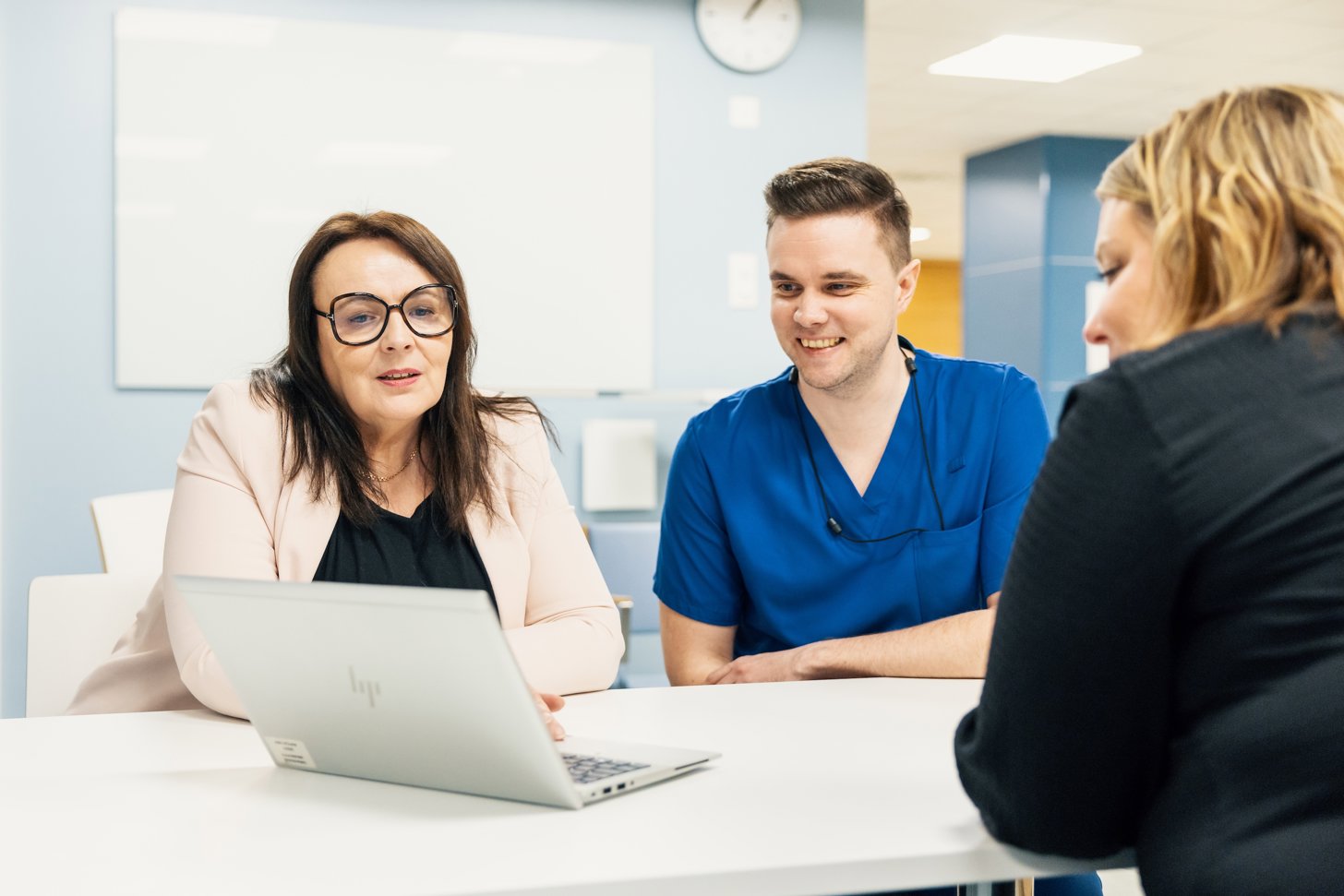 Three cheerful professionals sitting at a table looking at a laptop. From left: Marjo Patana (light pink blazer), Sami Penttilä (blue nurse shirt), and Tytti Valli (black jacket). In the background: light blue walls, a hallway, and a wall clock.